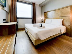 Bright hotel room at B&B Hotel Padova with a large bed, wooden headboard, folded towels, desk, chair, television, and window with brown curtains