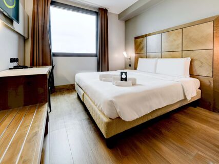 Bright hotel room at B&B Hotel Padova with a large bed, wooden headboard, folded towels, desk, chair, television, and window with brown curtains