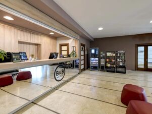 Reception area of B&B Hotel Padova with wooden front desk, computer monitors, vending machines, snack racks, and red stools