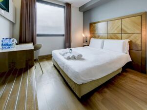 Modern hotel room at B&B Hotel Padova with a double bed, wooden headboard, rolled white towels, desk with water bottles, and a window with curtains