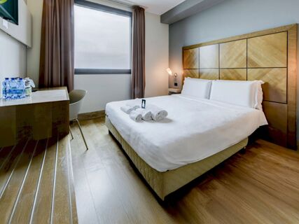 Modern hotel room at B&B Hotel Padova with a double bed, wooden headboard, rolled white towels, desk with water bottles, and a window with curtains