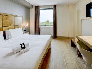 A bright hotel room at B&B Hotel Padova featuring a large bed with white linens, two rolled towels, a wall lamp, and a window with brown curtains showing trees outside
