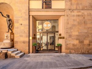 Entrance to B&B Hotel Firenze Pitti Palace al Ponte Vecchio with glass doors, two potted plants, and a bronze statue nearby