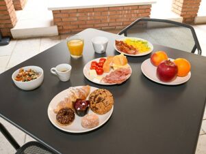 A breakfast spread with pastries, fresh fruit, cold cuts, scrambled eggs, orange juice, coffee, and muesli on a black table at B&B Hotel Affi Lago di Garda