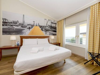 Double bed with white linens and towels, wooden floor, two bedside tables with lamps, large window, artwork with boats in B&B Hotel Affi Lago di Garda