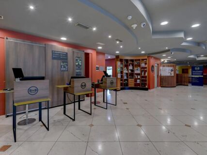 Spacious hotel lobby with three check-in kiosks, wooden shelving, informational signage, and tiled floor at B&B Hotel Affi Lago di Garda