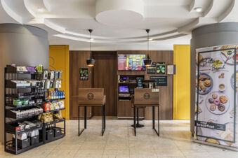 Lobby area of B&B Hotel Modena with two wooden check-in counters, snack racks, and a breakfast promotional banner