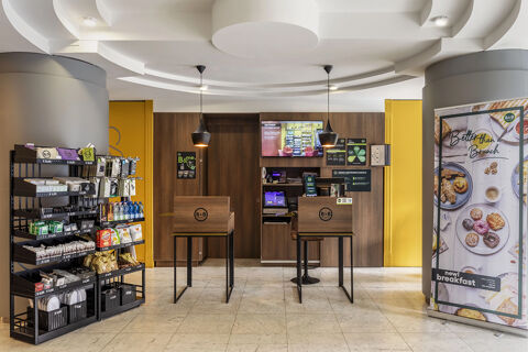 Lobby area of B&B Hotel Modena with two wooden check-in counters, snack racks, and a breakfast promotional banner