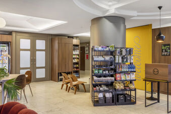 Interior lobby area of B&B Hotel Modena featuring snack shelves, modern chairs, a welcome sign on a yellow door, and a small reception desk