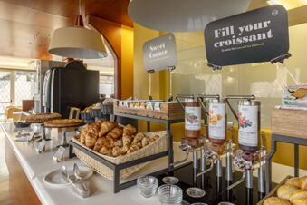 Buffet table at B&B Hotel Modena with a basket of croissants, jars of jam, coffee machine, cakes, and packaged snacks