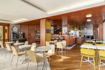 Dining area with round tables, wooden and yellow chairs, coffee machine, and serving counter inside B&B Hotel Modena