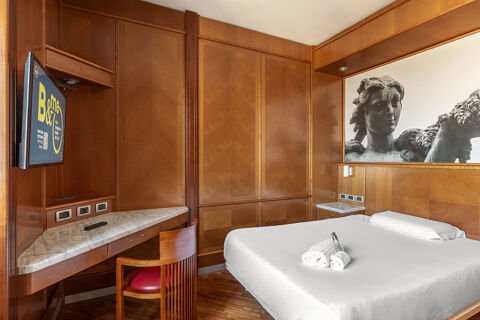 Hotel room at B&B Hotel Modena with wooden walls, a bed with white linens, a desk with a chair, and a TV mounted on the wall