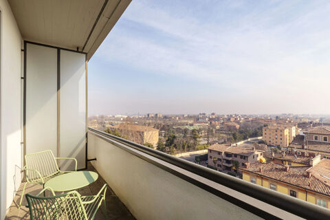 Balcony at B&B Hotel Modena with two green metal chairs and a small table overlooking city buildings and trees under a blue sky