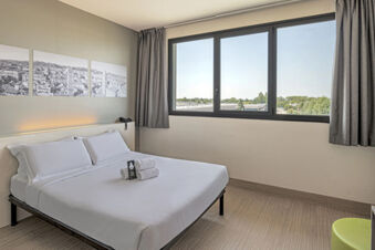 Modern hotel room at B&B Hotel Bologna with a neatly made bed, rolled towels, large window, and panoramic city photos on the wall