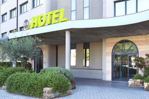 Entrance area of B&B Hotel Bologna with green bushes, stone planters, and a yellow hotel sign on the building facade