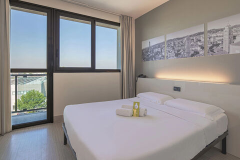 Minimalist hotel room at B&B Hotel Bologna with a double bed, rolled towels, a cityscape artwork, and windows with a view
