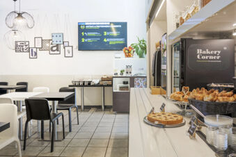 Bakery Corner with croissants, cake, and yogurt on a counter, tables, chairs, and a TV screen in B&B Hotel Bologna dining area