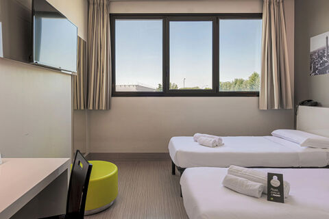 A hotel room at B&B Hotel Bologna with two single beds, a wall-mounted TV, a desk, and a large window with curtains