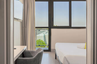 Minimalist hotel room at B&B Hotel Bologna with a bed, light gray armchair, white desk, and large window with curtains