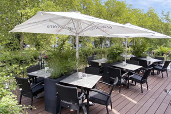 Terrasse mit Tischen, St&uuml;hlen und Sonnenschirmen umgeben von gr&uuml;nen Pflanzen beim Hotel am Westend an einem sonnigen Tag.