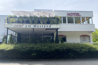Die Au&szlig;enansicht des Hotel am Westend zeigt ein modernes Geb&auml;ude mit einer Terrasse, Sonnenschirmen und viel Gr&uuml;n vor dem Eingangsbereich.