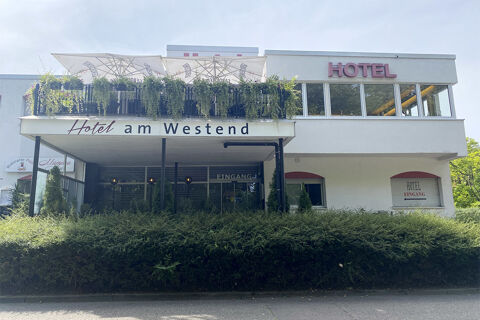 Die Au&szlig;enansicht des Hotel am Westend zeigt ein modernes Geb&auml;ude mit einer Terrasse, Sonnenschirmen und viel Gr&uuml;n vor dem Eingangsbereich.