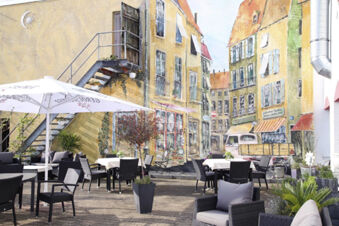 Sitzbereich im Freien mit Tischen, St&uuml;hlen und Sonnenschirmen vor einer farbenfrohen Wandbemalung beim Hotel am Westend.