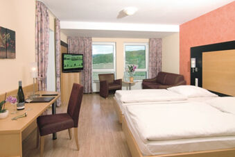 A hotel room at Burg-Hotel Obermoschel with two single beds, a sofa, armchair, desk with chair, and windows with curtains showing a green landscape view