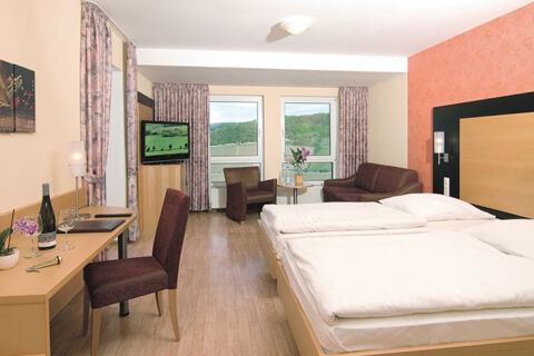 A hotel room at Burg-Hotel Obermoschel with two single beds, a sofa, armchair, desk with chair, and windows with curtains showing a green landscape view