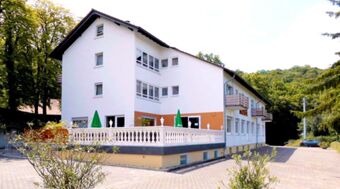 Large white building with three floors, balconies, green umbrellas on a terrace, and surrounding greenery at Burg-Hotel Obermoschel
