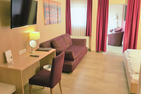 Hotel room at Burg-Hotel Obermoschel with a purple sofa, desk with chair and lamp, flat screen TV, and wooden flooring