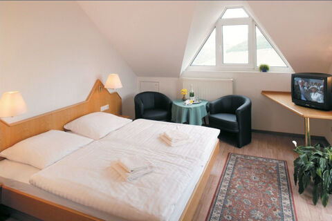 Double bed with white linens and towels, two black chairs, a table, a television, and a triangular window at Burg-Hotel Obermoschel
