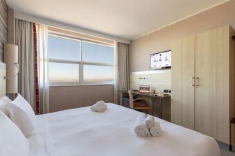 Bedroom interior at B&B Hotel Cherasco Langhe with a bed, rolled towels, large window, desk, chair, TV, and wardrobe visible