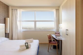 Modern hotel room at B&B Hotel Cherasco Langhe with a bed, towels, large window, desk, chair, and wardrobe visible inside