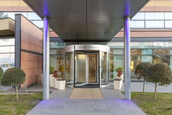 Entrance of B&B Hotel Cherasco Langhe with glass revolving door, beige tiles, two silver pillars, and potted plants on sides