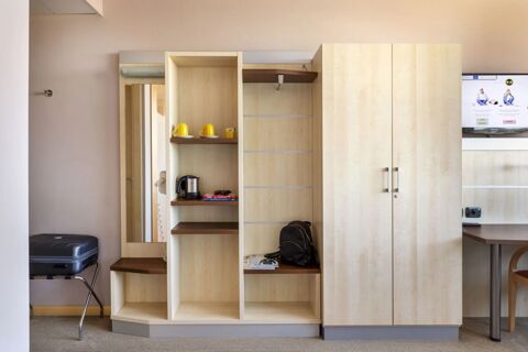 Light wood shelving unit with yellow mugs, electric kettle, sunglasses, books, black backpack, wardrobe, and a Samsung TV at B&B Hotel Cherasco Langhe
