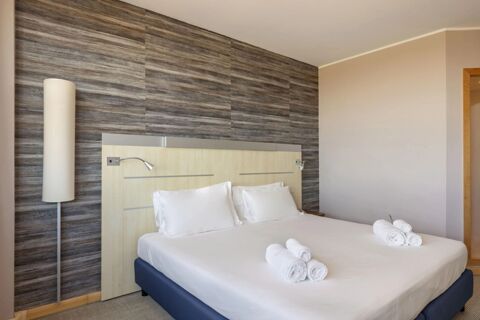 Bed with white linens and pillows, a wooden headboard, rolled towels, and a tall floor lamp at B&B Hotel Cherasco Langhe