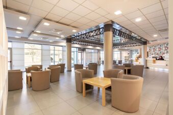 Lobby of B&B Hotel Cherasco Langhe with beige armchairs, wooden tables, large windows, and reception desk with staff talking on phone