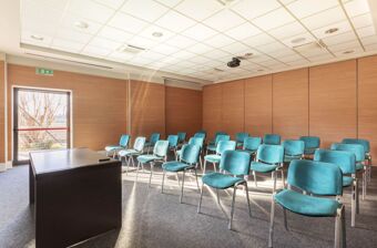 Conference room with blue chairs, a wooden podium, carpeted floor, wood-paneled walls, and a glass door at B&B Hotel Cherasco Langhe