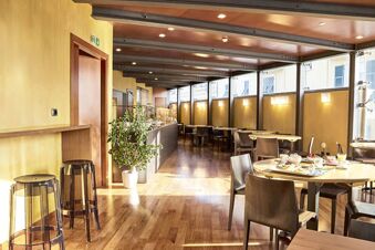 Dining area of B&B Hotel Imperia Rossini al Teatro with wooden tables, chairs, glass stools, and natural lighting from windows