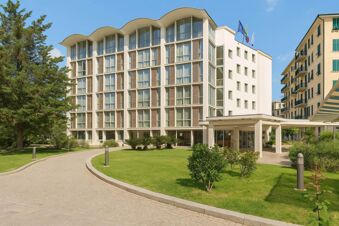 Modern multi-story building with large windows and a curved roof next to a curved walkway and green lawns at B&B Hotel Imperia Rossini al Teatro