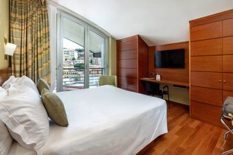 Double bed with white linens and green pillows next to large windows with balcony and wooden furniture at B&B Hotel Imperia Rossini al Teatro