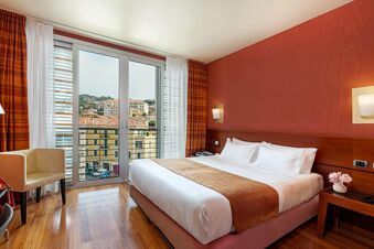 Bedroom at B&B Hotel Imperia Rossini al Teatro featuring a large made bed, wooden floor, beige armchair, and glass door to balcony view