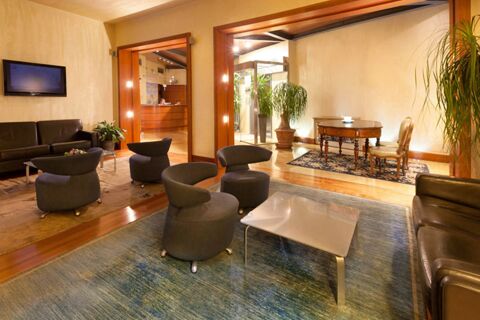 Lobby area of B&B Hotel Imperia Rossini al Teatro with multiple gray chairs, brown leather sofas, a TV, and potted plants