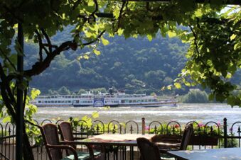 Outdoor seating with tables and chairs under grapevines overlooking a river with a paddle steamer boat near Rheinhotel Zum Anker