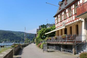Riverside view of Rheinhotel Zum Anker with flowers on balcony, a stone walkway, and hills under a clear sky
