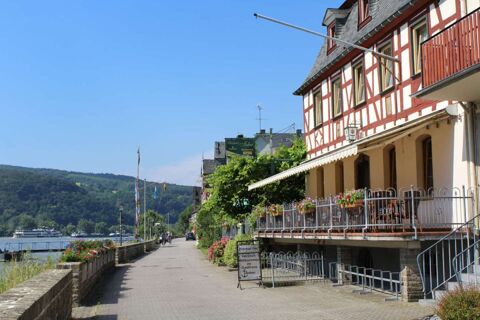 Riverside view of Rheinhotel Zum Anker with flowers on balcony, a stone walkway, and hills under a clear sky