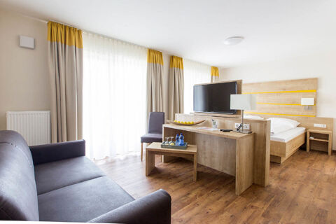 Modern hotel room at Landgasthof Bieg with a bed, wooden furniture, TV, gray sofa, chair, and large windows with curtains