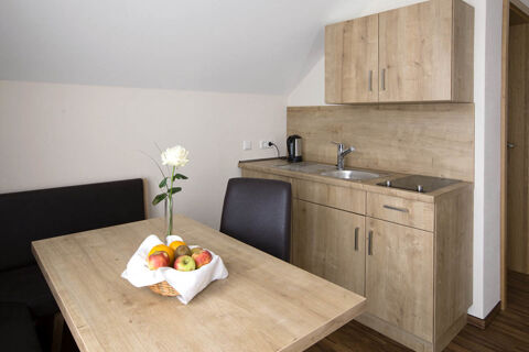 Small kitchen area with wooden cabinets, sink, electric stovetop, and table with chair and fruit basket at Landgasthof Bieg
