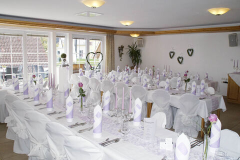 A bright banquet room at Landgasthof Bieg with white-covered chairs, lilac and white folded napkins, pink candles, and floral decorations